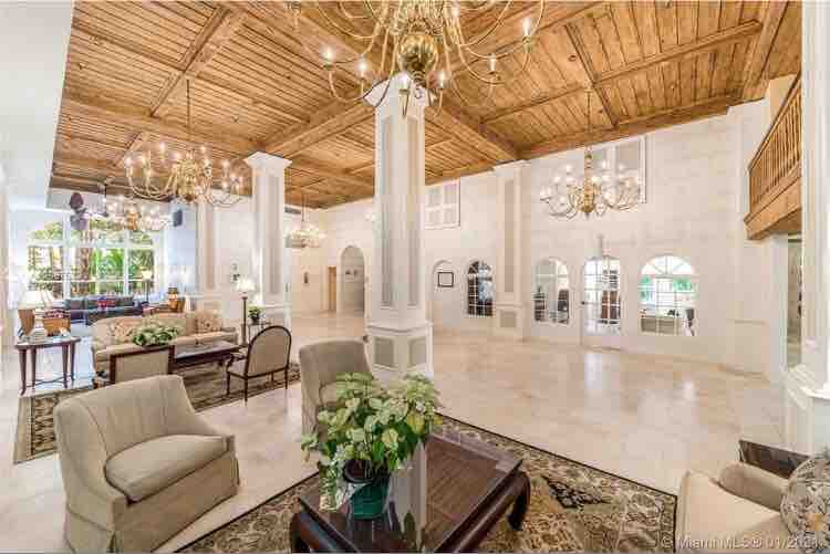 A spacious lobby area is characterized by high ceilings with wooden beams and elegant chandeliers. Light-colored stone floors complement the cream-toned walls. Comfortable seating arrangements are present, accompanied by decorative plants, creating an open and inviting environment.