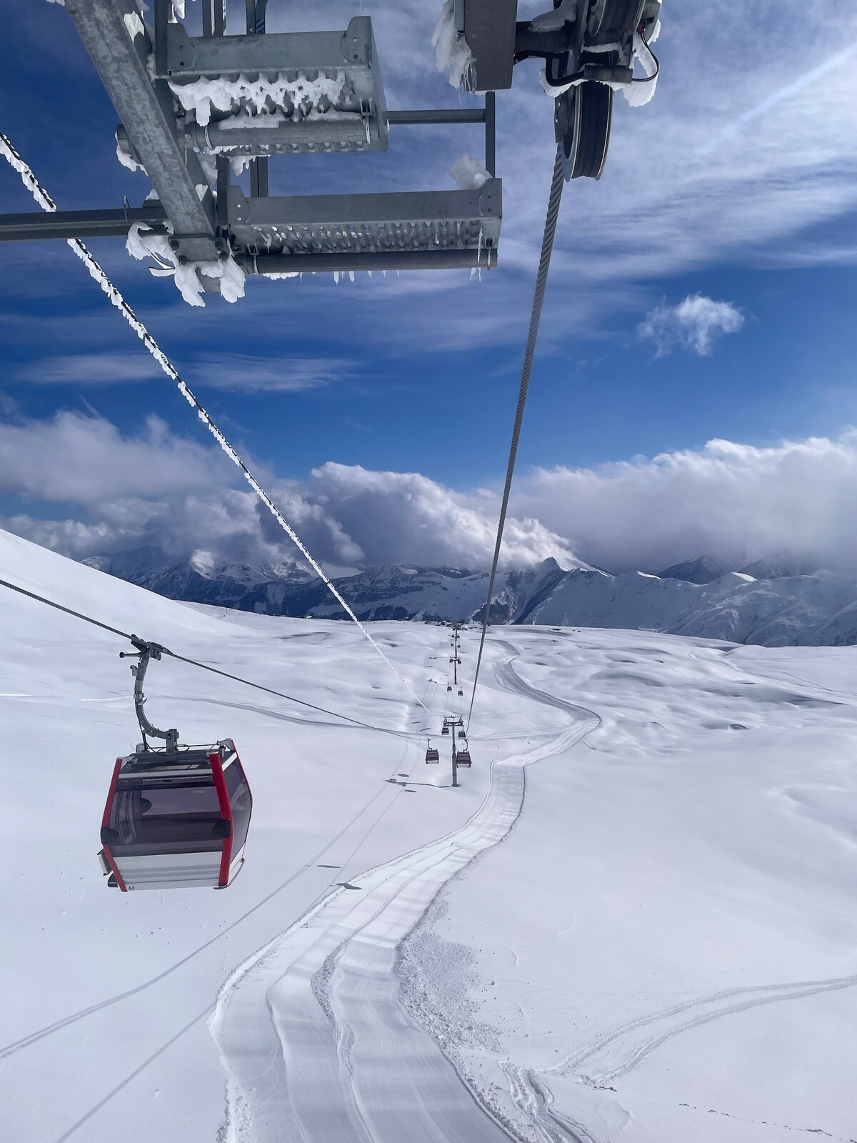 A snow-covered landscape extends beneath a ski lift, showing a series of gondolas ascending above the pristine white terrain. Mountains rise in the background, partially obscured by fluffy clouds under a clear blue sky.
