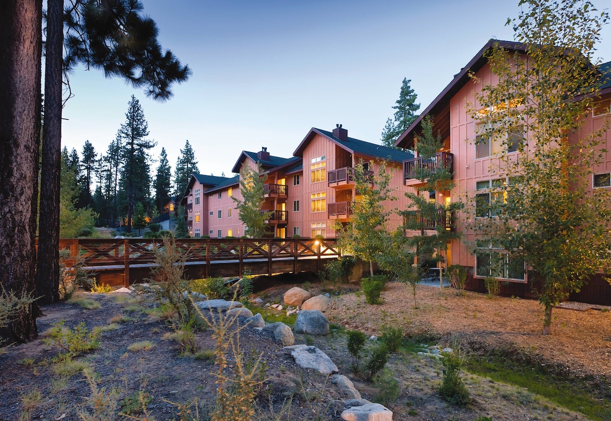 A resort building stands amidst tall trees, illuminated gently in the evening light. A wooden bridge crosses a tranquil area, enhancing the natural surroundings. Landscaping features shrubs and stones, creating a serene atmosphere. The architecture reflects a cozy design, suggesting a welcoming retreat.