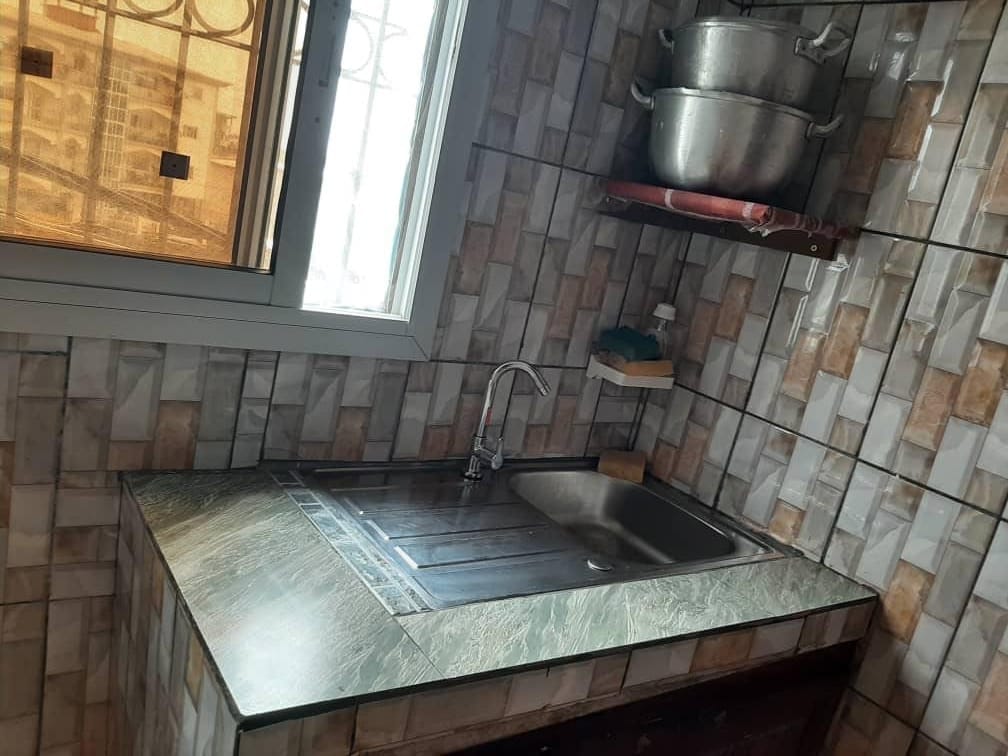 A compact kitchen area features a stainless steel sink set into a marble countertop. Natural light illuminates the space through a window with a barred grill. Metallic pots are stored on an overhead shelf, with beige and brown patterned tiles covering the walls.