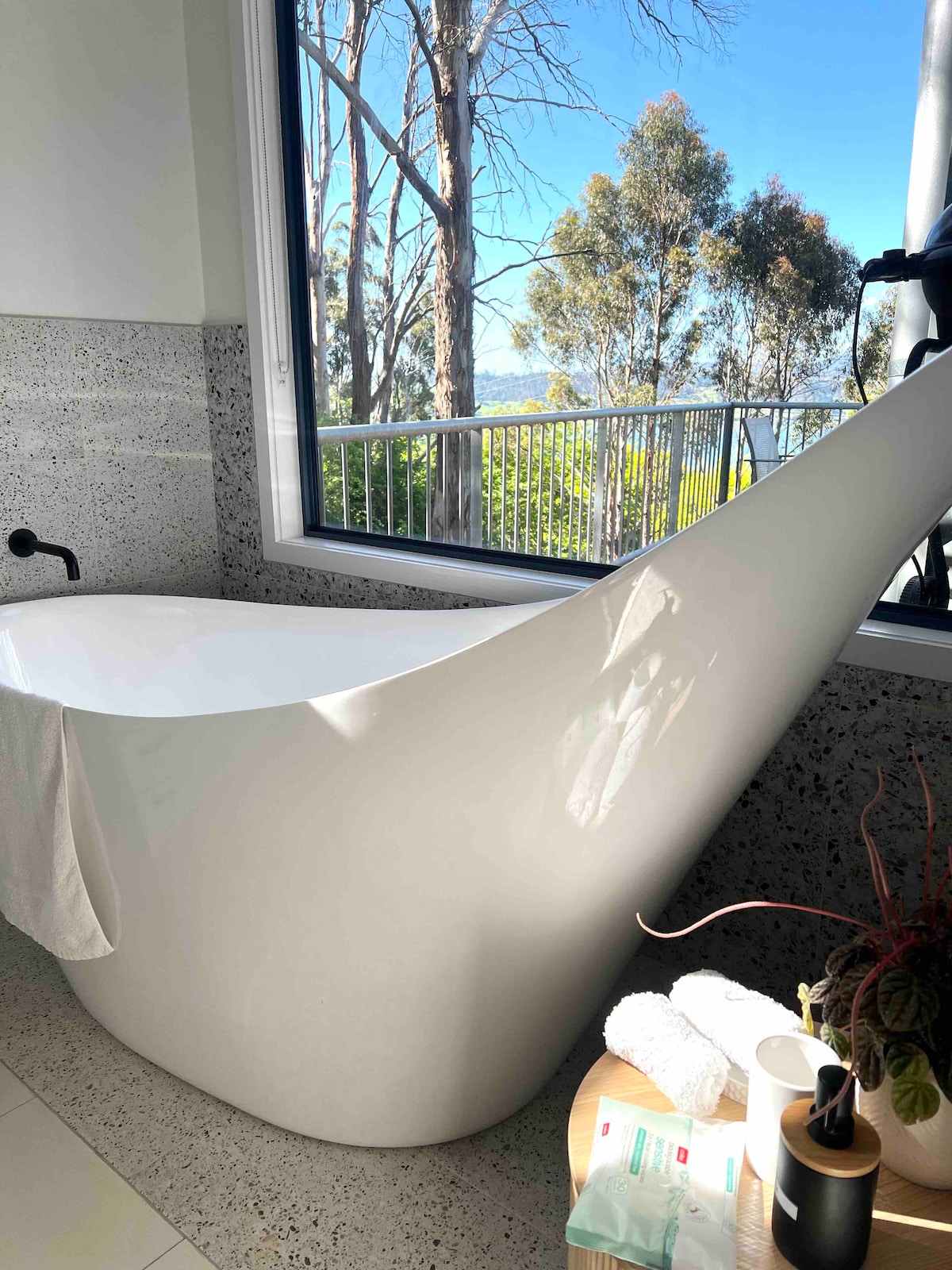A freestanding bathtub is positioned near a large window, offering views of the surrounding trees and outdoor space. Natural light fills the bathroom, highlighting the textured wall and modern design. A plant and neatly arranged towels add subtle detail to the serene setting.