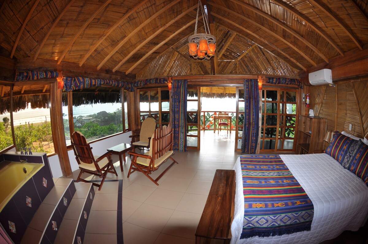 A spacious room is presented with a thatched roof, featuring large glass windows that open to a scenic view. A comfortable bed with a colorful blanket is positioned near a wooden bench, with seating arrangements including a pair of rocking chairs and a small table.