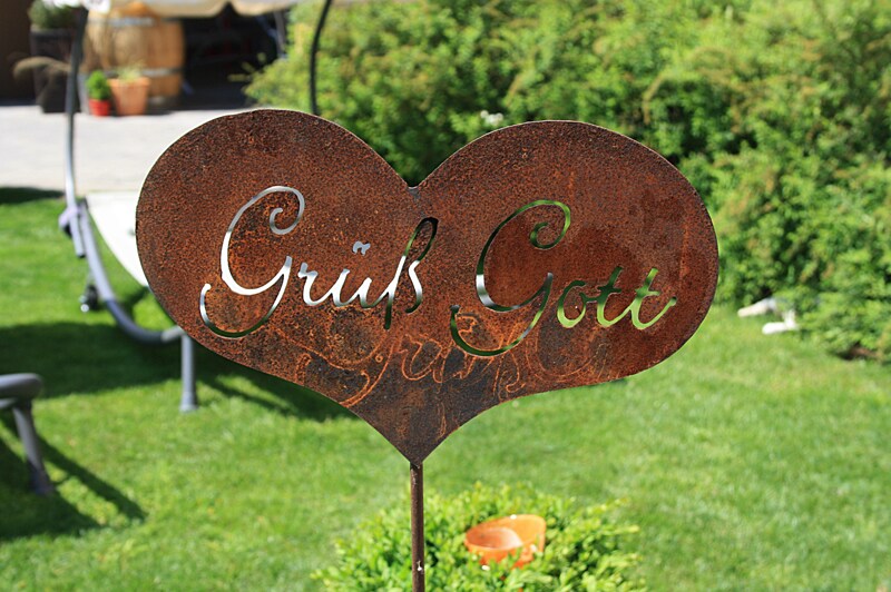 A rusted metal sign in the shape of a heart is seen against a backdrop of lush greenery. The sign features the greeting 'Grüß Gott' cut out in an elegant script, adding a welcoming touch to the outdoor space.
