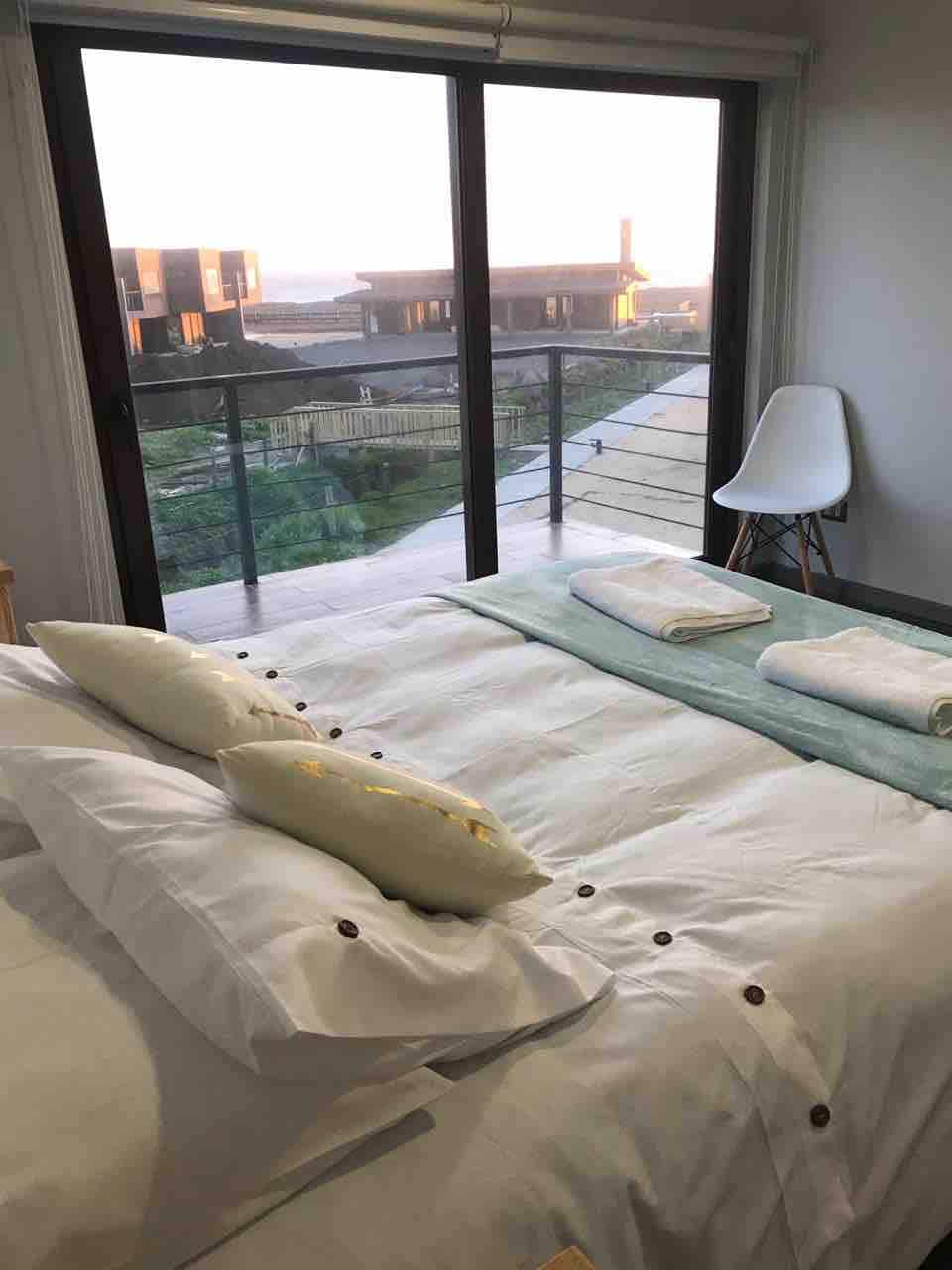 A serene sleeping area is furnished with a spacious bed adorned with plush white linens and decorative pillows. Two rolled towels are placed neatly on the bed. Large glass doors provide a view of the outdoor area and natural surroundings, allowing ample sunlight to brighten the room.