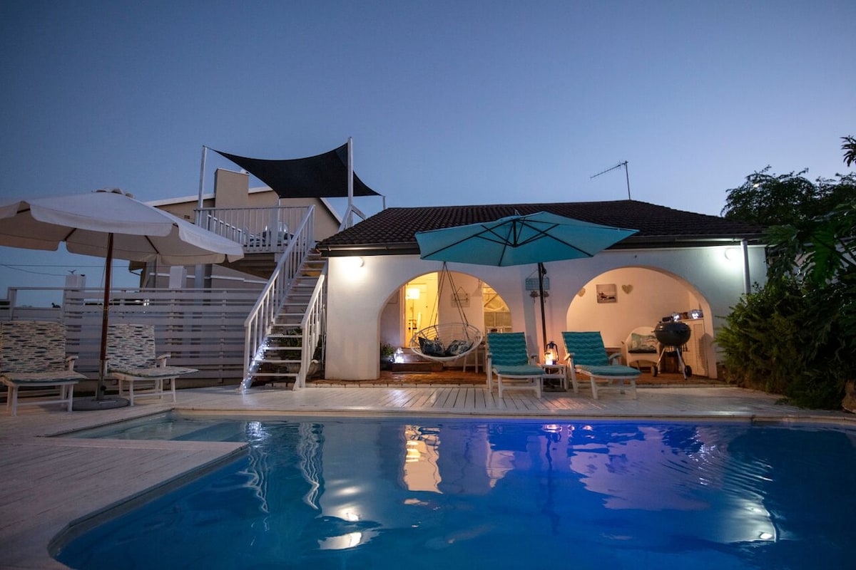 An outdoor area features a swimming pool reflecting twilight hues, surrounded by loungers and an umbrella. The structure offers an arched entrance leading into a well-lit interior. A staircase is visible, along with a hammock for relaxation, enhancing the recreational space.
