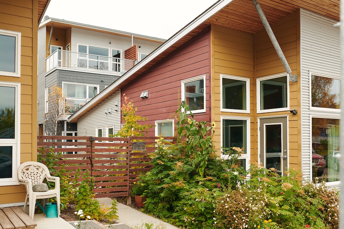 A modern multi-story building is depicted, featuring a mix of brown and yellow paneling. Lush greenery and various flowers surround the entrance, complemented by a small patio area with a single chair. Large windows allow natural light to illuminate the interior.