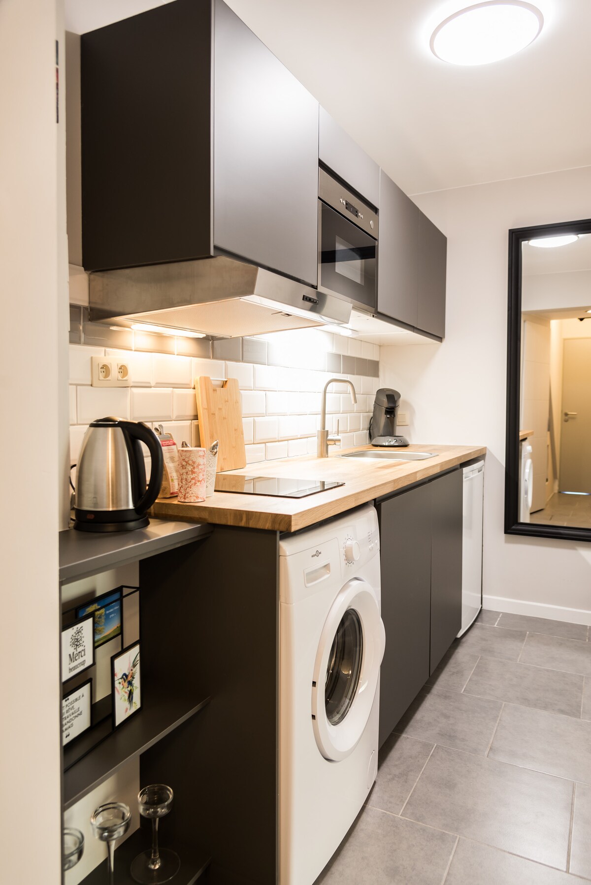 A modern kitchen area is visible, featuring sleek black cabinetry and a wooden countertop. Appliances include a kettle, multi-function microwave, and a washing machine. A large mirror reflects the space, enhancing its openness, while subtle lighting ensures a well-lit and functional cooking environment.