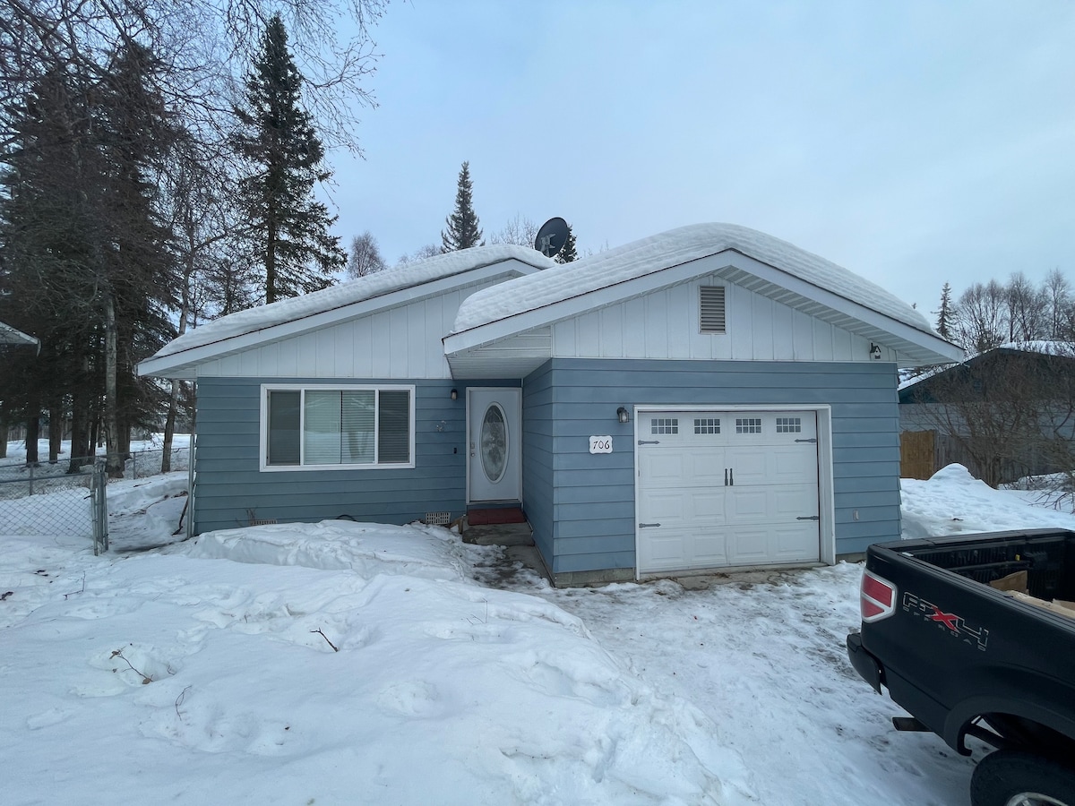 A light blue single-story house features a garage and a front door with a decorative circular window. Snow covers the driveway and yard, while evergreen trees are visible in the background. The roof is newly coated in white snow, creating a uniform winter scene.