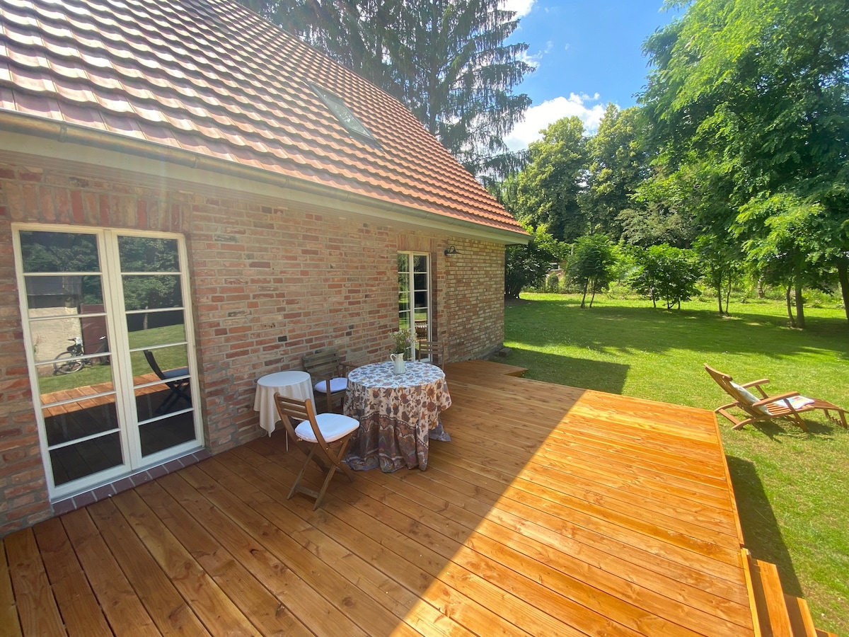 An outdoor terrace features a wooden deck with a round table set for dining, surrounded by trees and grassy areas. Wooden deck chairs are positioned nearby, inviting relaxation in a serene setting with ample sunlight.