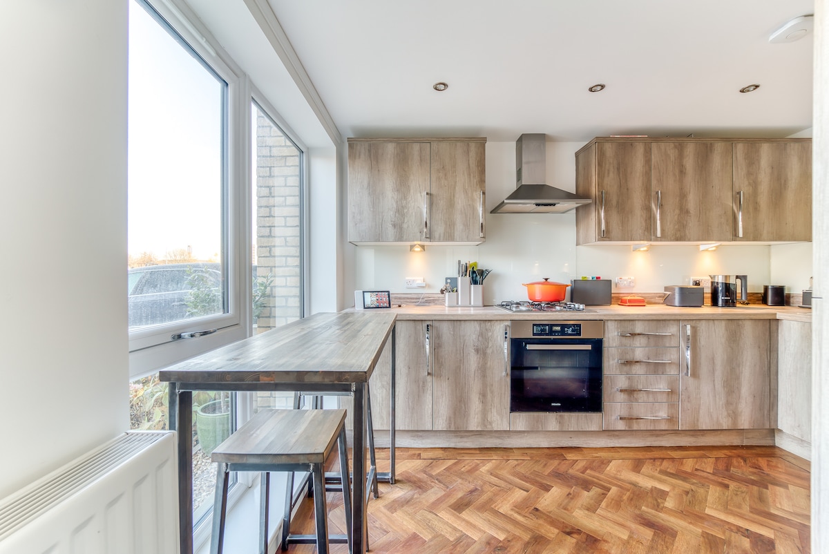 A modern kitchen features wooden cabinetry with stainless steel appliances. A wooden dining table with two stools stands beside a large window, allowing natural light to fill the space. Under-cabinet lighting highlights the countertop, while the oven and stove are positioned centrally.
