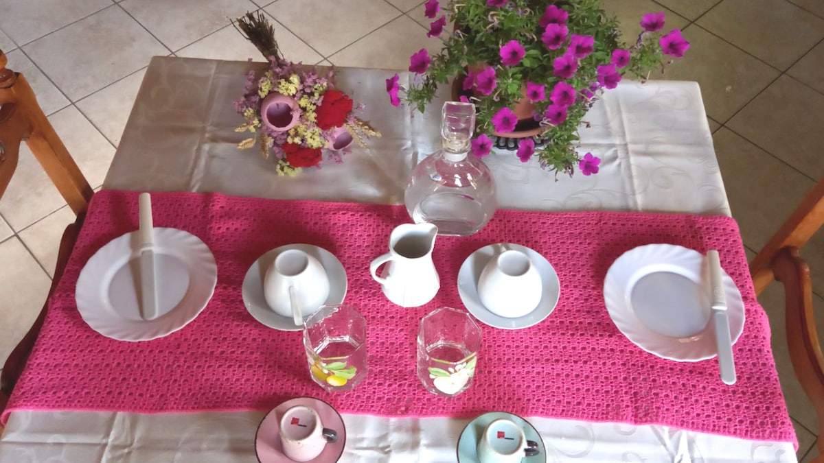 A dining table is set with four white plates and paired with glasses, a pitcher, and a small cream jug. A pink crochet runner adds color, complemented by a floral centerpiece with vibrant flowers. Understated elegance is conveyed in the arrangement, enhancing an inviting dining experience.