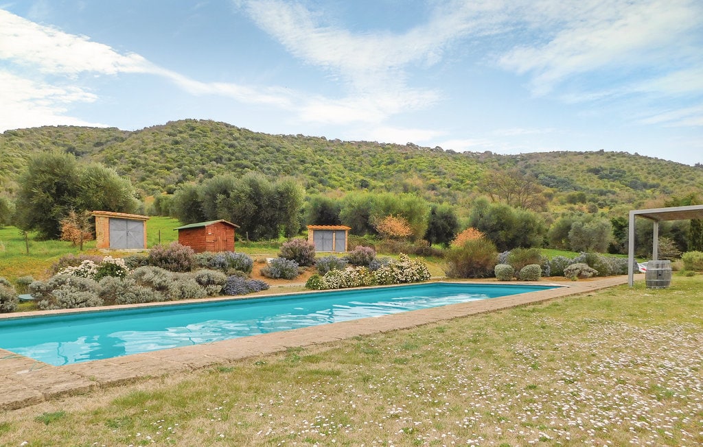 An outdoor swimming pool is set against a backdrop of rolling hills and greenery, surrounded by well-maintained flower beds. Several wooden structures are positioned nearby, with olive trees dotting the landscape. The sky is partly cloudy, enhancing the serene atmosphere of the countryside.