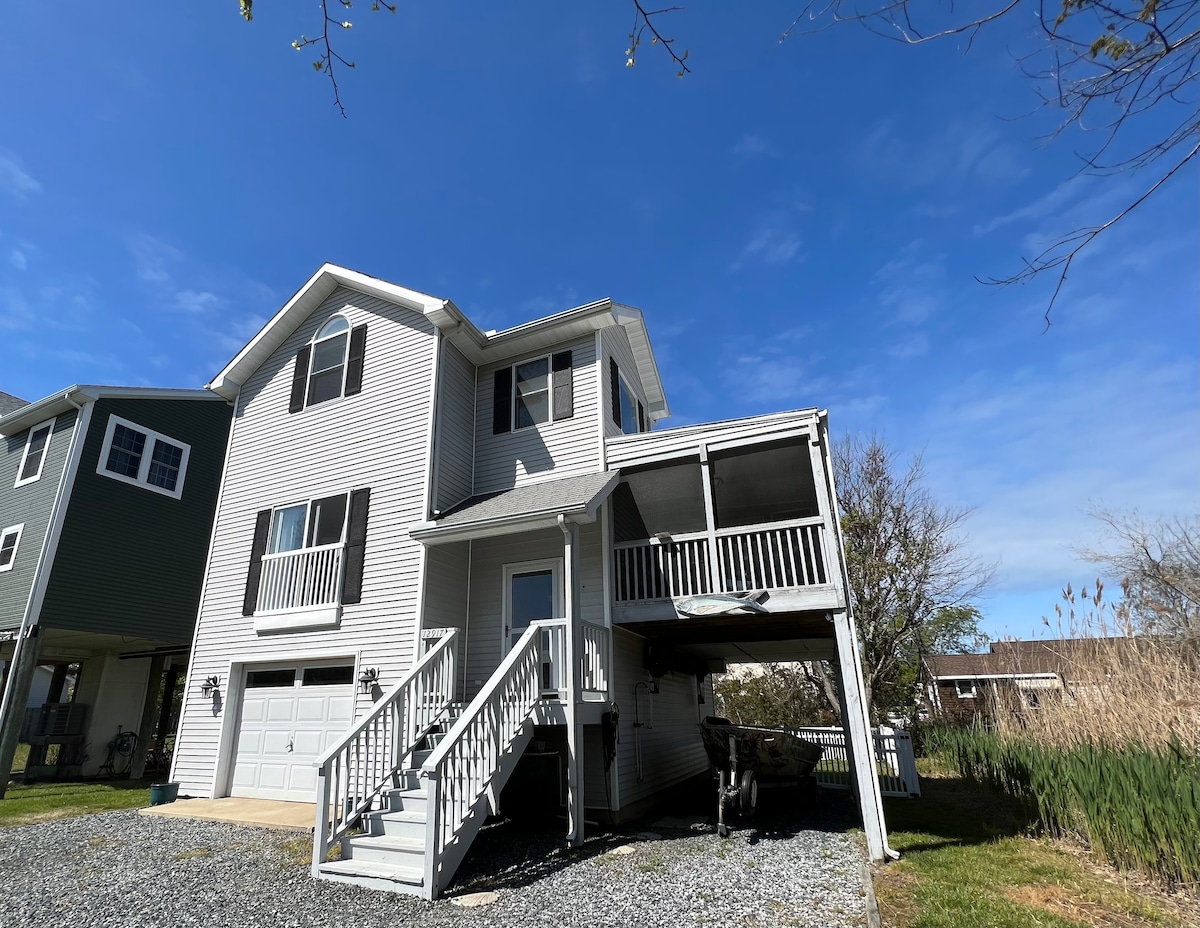 The exterior of a two-story house is presented, featuring a covered porch with steps leading to the entrance. A garage is visible on the ground level, with a gravel driveway. The surrounding area includes tall grasses and a clear blue sky.