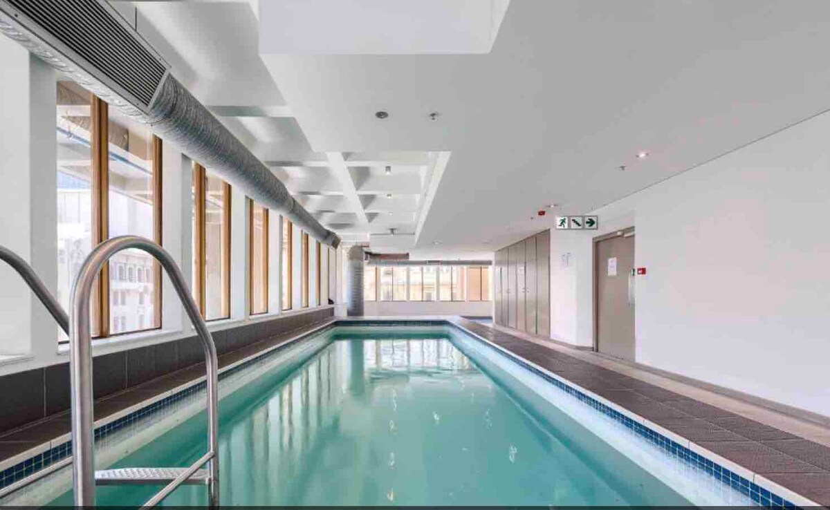 An indoor pool area is illuminated by natural light filtering through large windows. The pool, lined with tiles, is flanked by a sleek deck and features a stainless steel ladder. Neutral tones and modern design elements create a clean and inviting environment.
