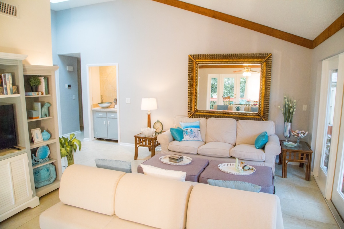The spacious living area features light cream sofas arranged around a central ottoman. A large mirror reflects the natural light, while a wooden beam accentuates the vaulted ceiling. Nearby, glass doors lead to an outdoor space, adding an openness to the room.