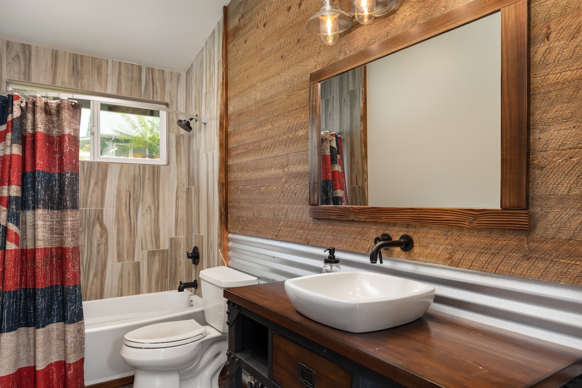 A modern bathroom features wood paneling and a contrasting metal accent wall. A spacious vanity with a sleek vessel sink is complemented by a large mirror. The bathtub is surrounded by stylish tiles, and a colorful shower curtain adds a touch of character.