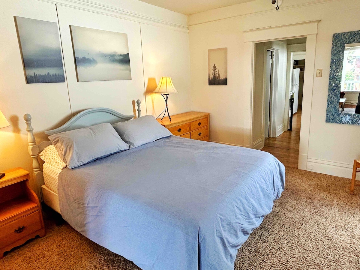 A spacious bedroom is highlighted by a queen-size bed with light blue bedding, positioned between two lamps. Framed landscape images adorn the walls, while a wooden dresser provides storage. Soft carpet underfoot and an open doorway lead to other areas of the apartment.