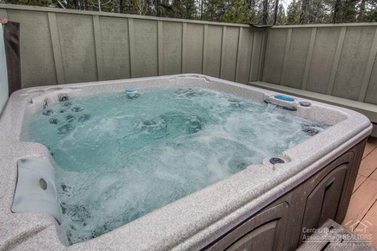 An outdoor hot tub is set on a wooden deck, featuring bubbling water and built-in jets. The surrounding area includes a privacy barrier, with trees visible in the background, suggesting a tranquil atmosphere for relaxation.