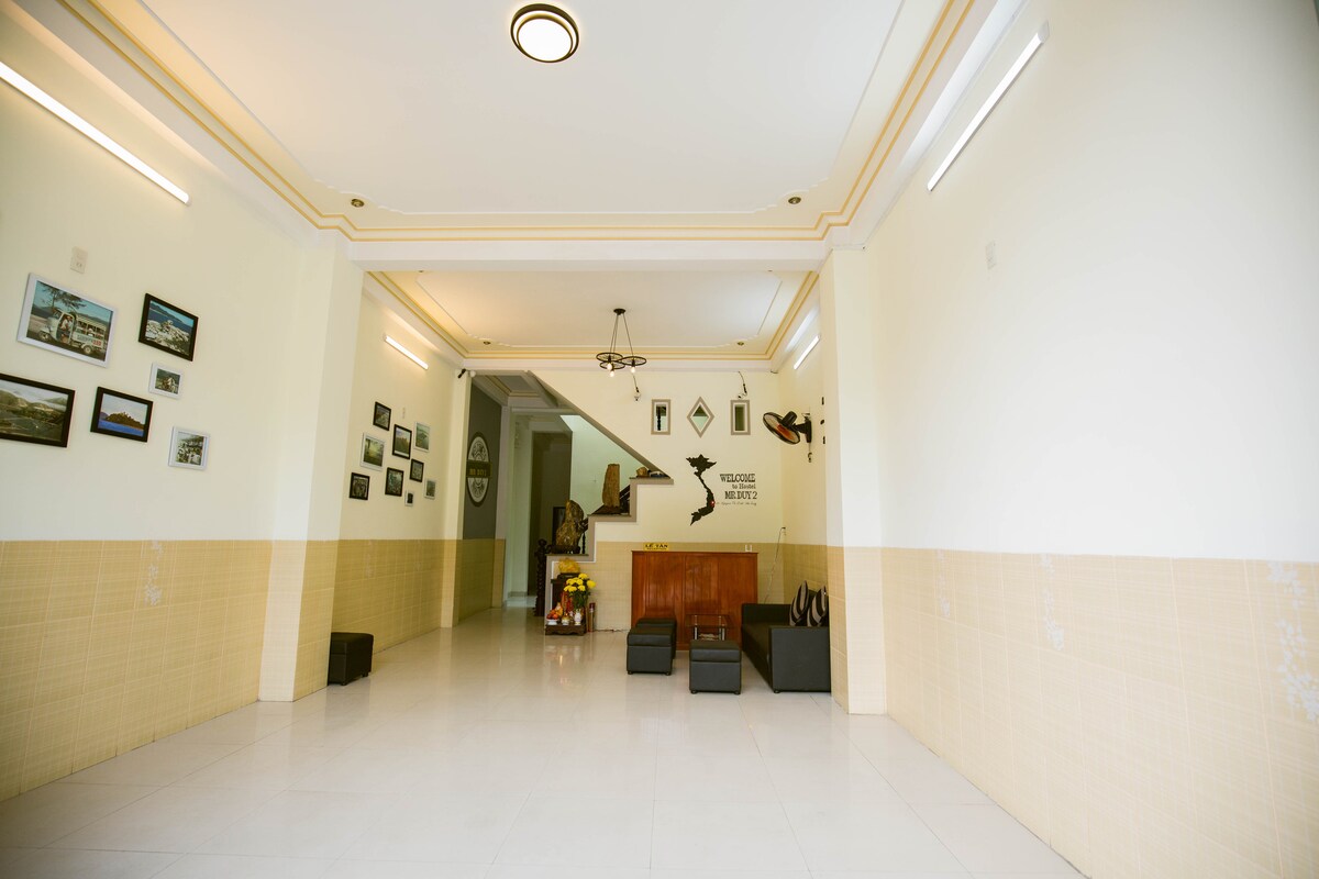 An open lobby area is visible, featuring sleek tile flooring and pale walls. Simple seating arrangements are provided with dark-colored furniture. The area is adorned with various framed photographs on the walls, and natural light enhances the spacious atmosphere.