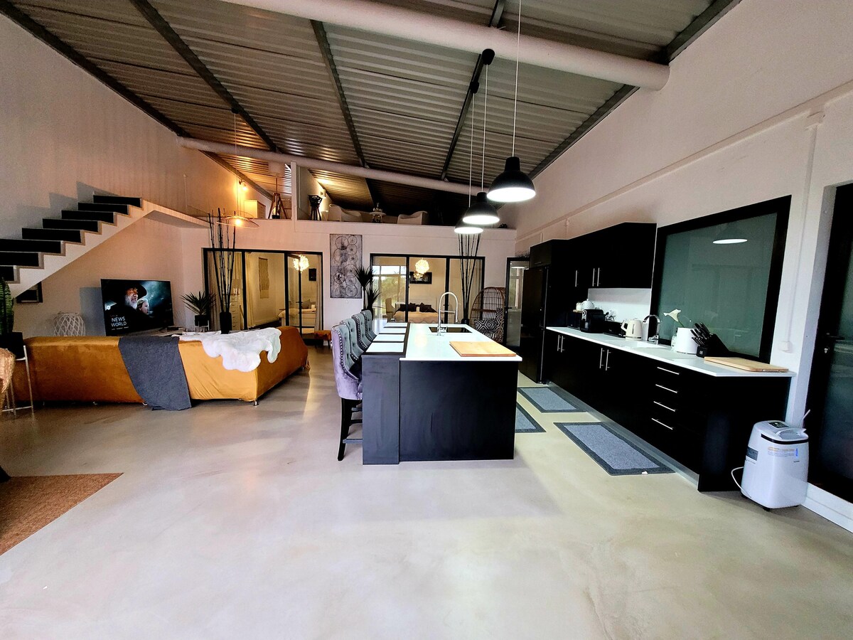 An open-concept living area showcases a modern kitchen with black cabinetry and an island, complemented by a spacious dining area. Light floods in through large windows, illuminating a cozy seating arrangement with a mustard-colored sofa, while a flight of stairs leads to an upper level.
