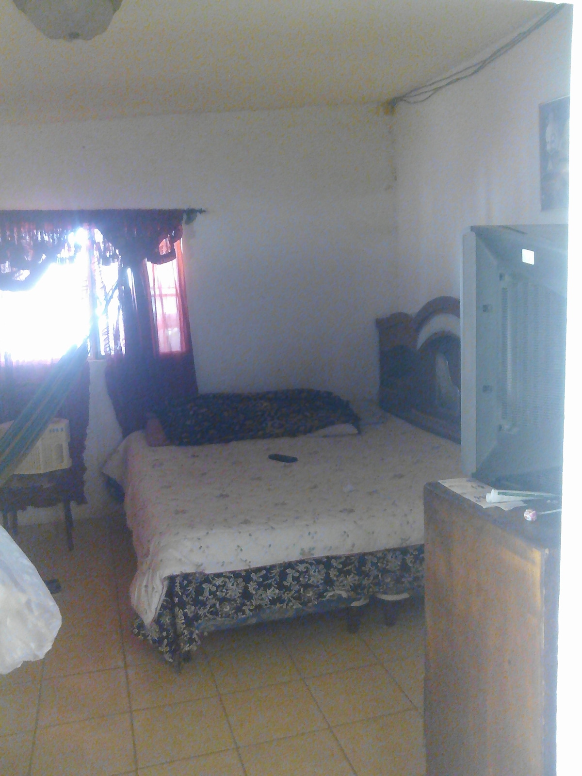 A cozy sleeping area features a spacious bed with a patterned cover. A television is placed on a wooden stand, while a window draped with colorful curtains allows natural light to enter the room. Light tiles complement the simple decor.