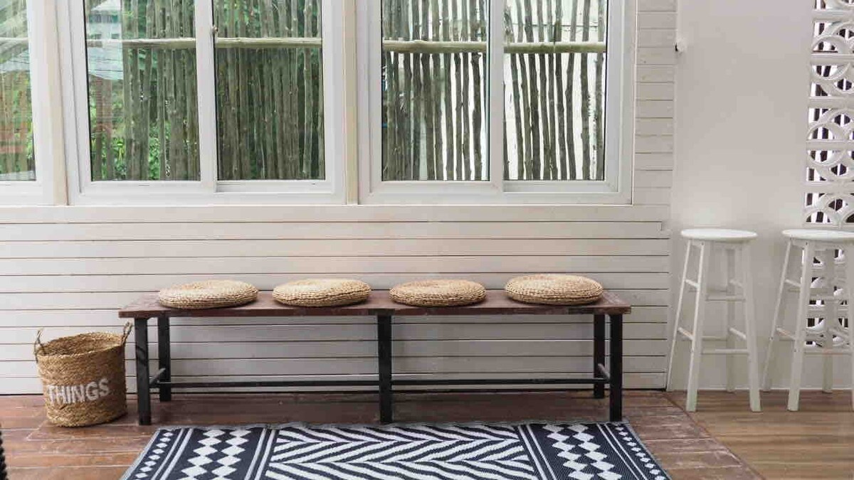 A seating area features a wooden bench with four round woven cushions. Adjacent to the bench, a small round table stands on one side, while large windows with bamboo blinds allow natural light to fill the space. A contemporary patterned rug lies underneath.