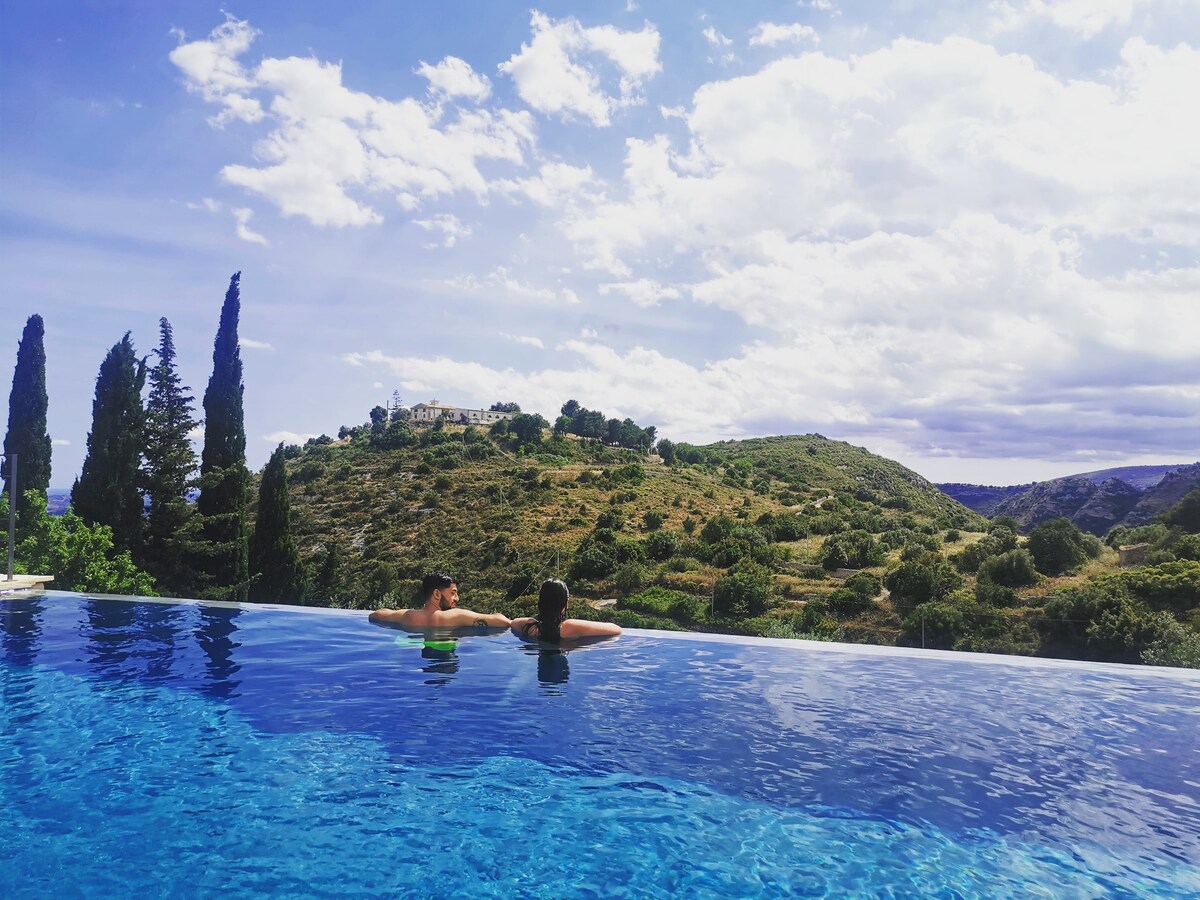 Individuals are seen relaxing in an infinity salt-water pool, overlooking lush green hills and distant trees. The sky is filled with fluffy clouds, and the surrounding landscape provides a serene backdrop.