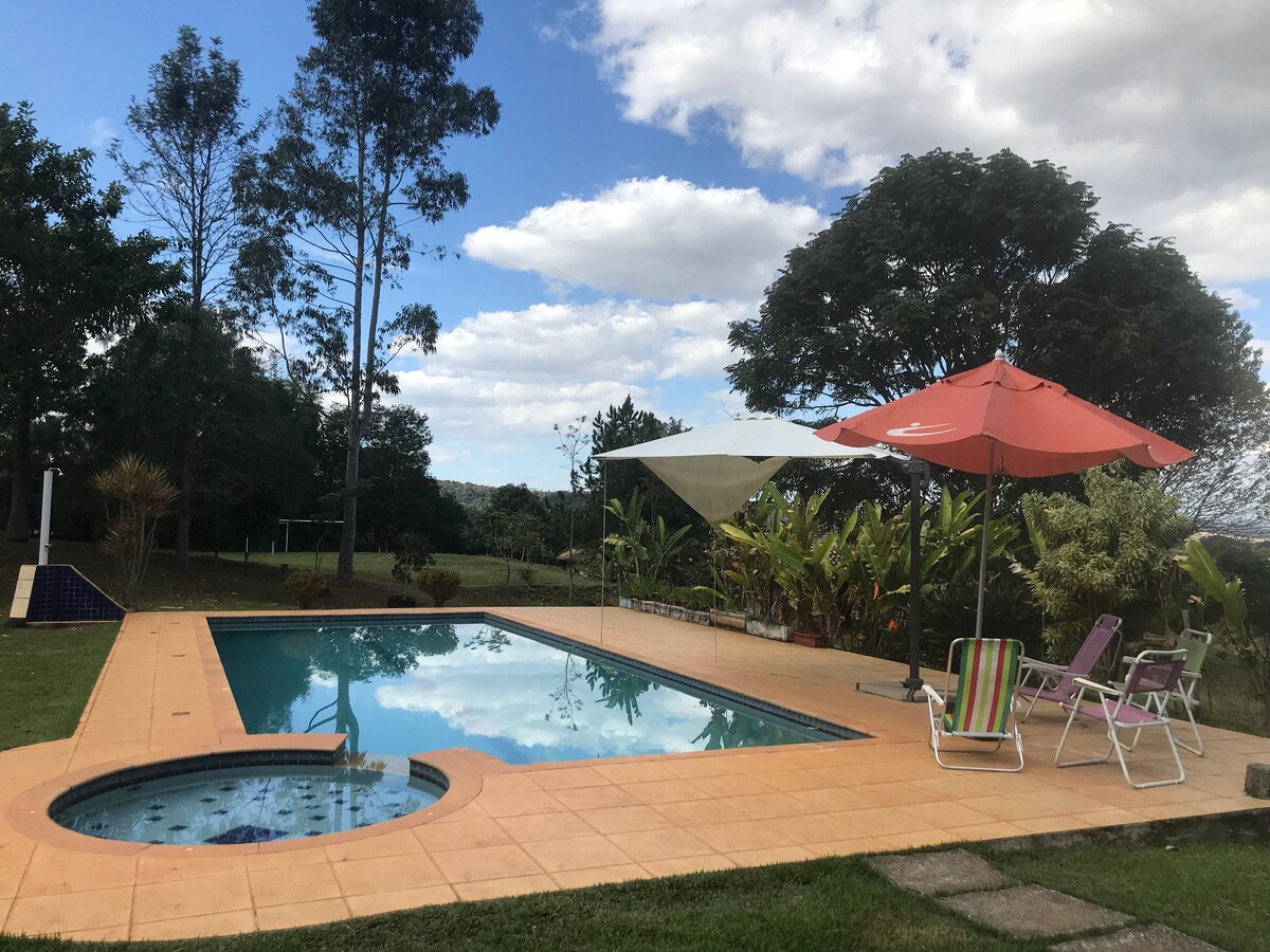 A tranquil outdoor area features a swimming pool surrounded by lush greenery and lounge chairs. Bright umbrellas provide shade, and the pool reflects the sky, while the landscape includes trees and open space, contributing to a serene environment.