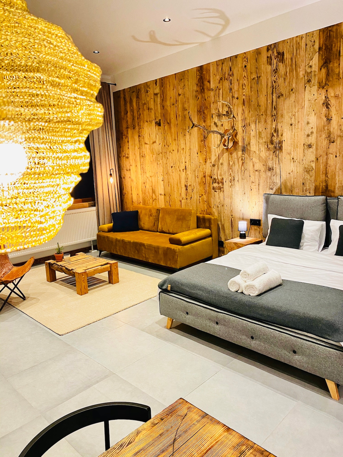 The interior features a spacious sleeping area with wood-paneled walls and soft, neutral tones. A cozy sofa and minimalistic coffee table complement a large bed, while an elaborate pendant light provides warm illumination. Towels are neatly arranged on the bed, enhancing the inviting atmosphere.