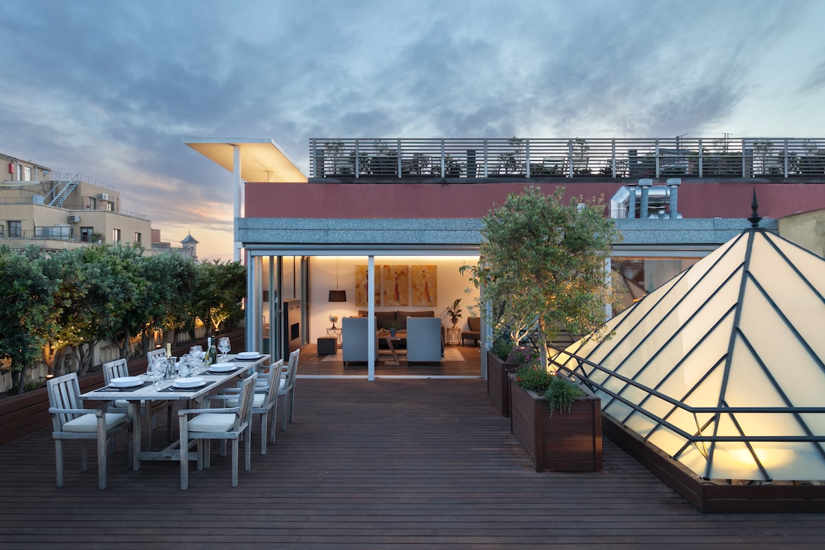 A spacious private terrace is showcased with a wooden deck and elegant outdoor dining set. Lush greenery surrounds the area, while a large glass structure adds architectural interest. The evening sky features soft clouds, and the interior space is partially visible through expansive glass doors.