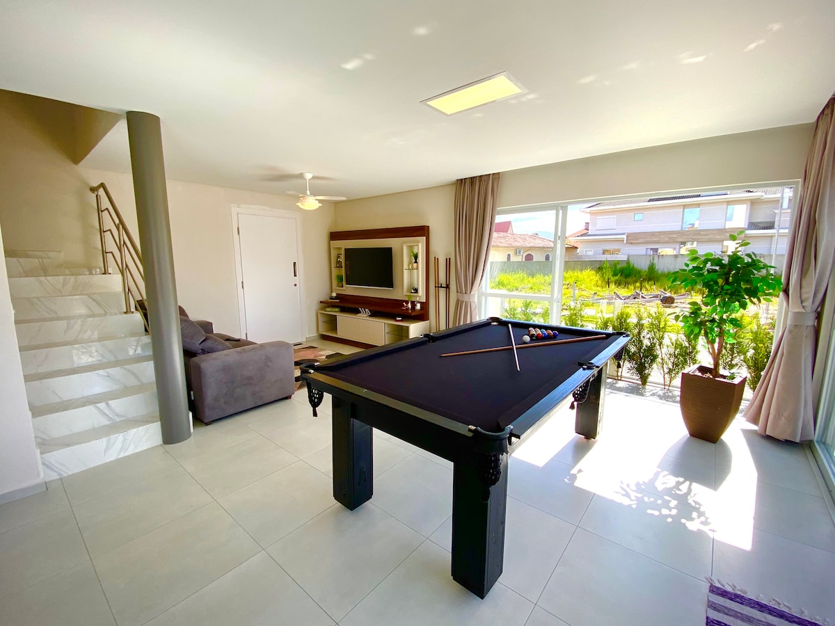 An inviting living area features a billiard table centrally placed, enhancing an atmosphere for leisure. Large windows allow natural light to illuminate the space, with a couch and entertainment center visible. Indoor plants add a touch of greenery, contributing to a relaxed environment.