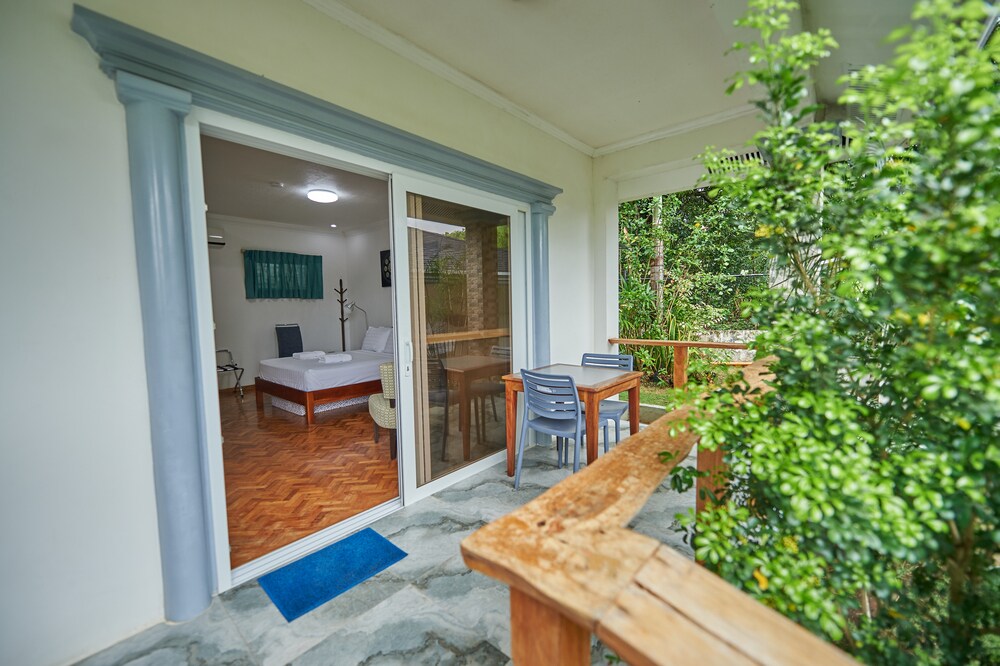 A private terrace is visible, featuring a wooden railing and outdoor seating. Through glass sliding doors, a bright interior is seen, showcasing a spacious bed, a small table with chairs, and greenery surrounding the entrance, indicating a serene atmosphere.