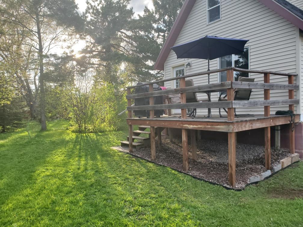 An exterior view of the house is shown, featuring a wooden deck with outdoor seating and an umbrella. Sunlight filters through the trees, casting shadows on the green lawn. The surrounding yard is landscaped with grass and shrubs.