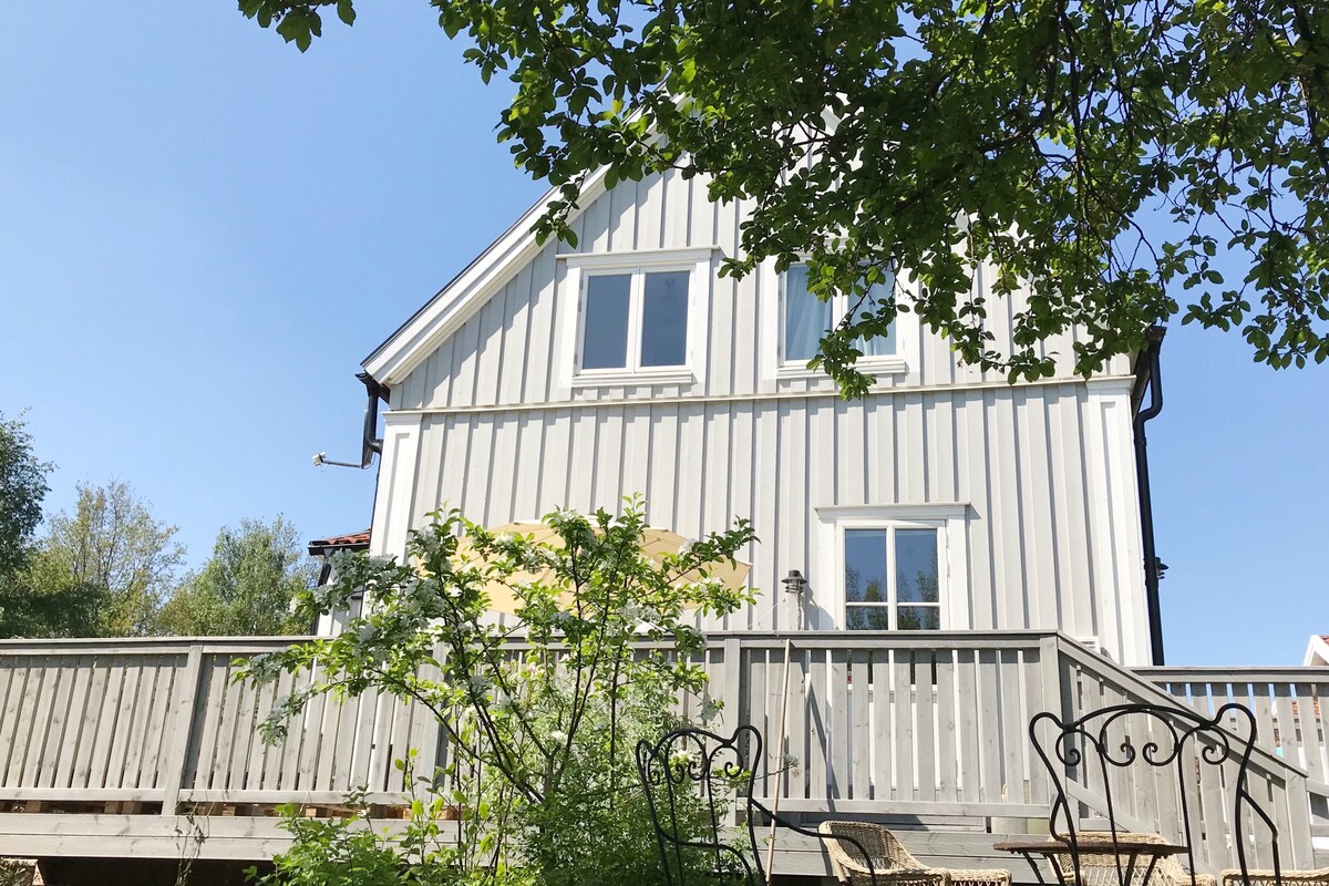The exterior of a charming two-story house is visible, featuring light-colored wooden siding and multiple windows. A wooden deck extends from the home, surrounded by greenery. A few simple outdoor chairs are positioned on the deck, inviting relaxation in the natural setting.