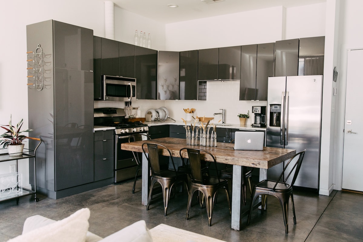 A modern kitchen is presented with sleek, dark cabinetry and stainless steel appliances. A wooden dining table with metal chairs is centrally situated, accompanied by decorative vases. Ample natural light illuminates the space, highlighting the contemporary design and open layout.
