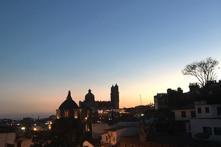A panoramic view of Taxco captures the evening sky transitioning from blue to orange, with silhouettes of iconic buildings including the church of Santa Prisca. Rooftops of nearby structures are visible, accompanied by distant trees framing the horizon.