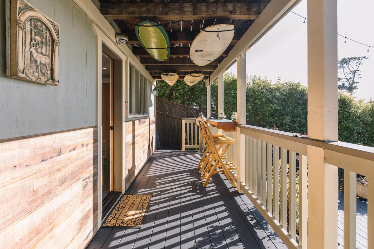 A covered porch area features seating at a high top table, with two chairs set against a backdrop of wood paneling. Surfboards are displayed on the ceiling, and a welcome mat is placed at the entrance to enhance the inviting atmosphere.