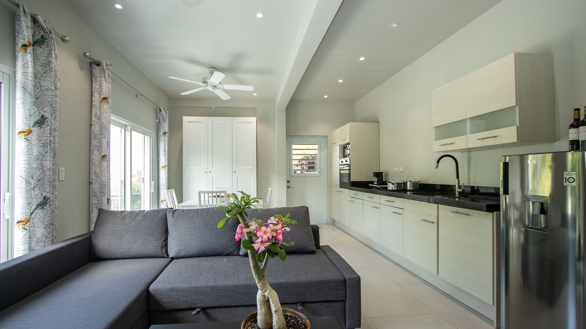 The open-plan living area features a modern sectional sofa positioned near large windows that allow natural light to fill the space. A dining table is visible in the background, accompanied by a well-equipped kitchen with sleek cabinetry and appliances.