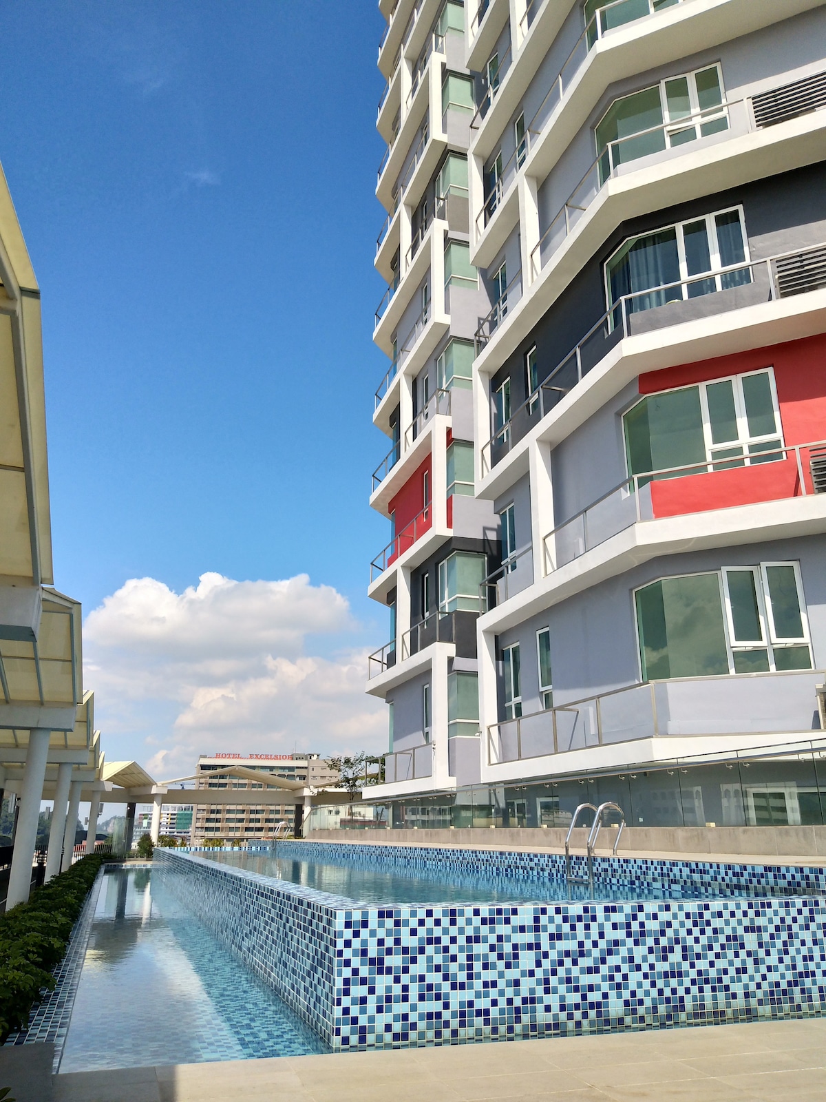 The image showcases a modern condominium structure with multiple floors. A long, rectangular swimming pool extends alongside the building, featuring a mosaic tile design. Clear blue skies complement the vibrant exterior colors of the building, creating a welcoming atmosphere for guests.