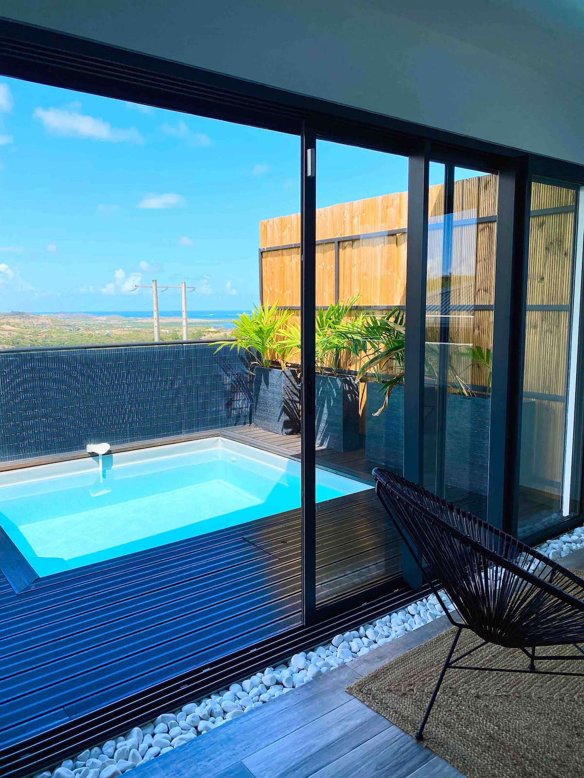 A private terrace displays a small pool, surrounded by well-maintained plants. Large glass doors invite natural light and showcase a view of the landscape. A black chair is positioned near the edge of the deck, adding a functional element to the space.