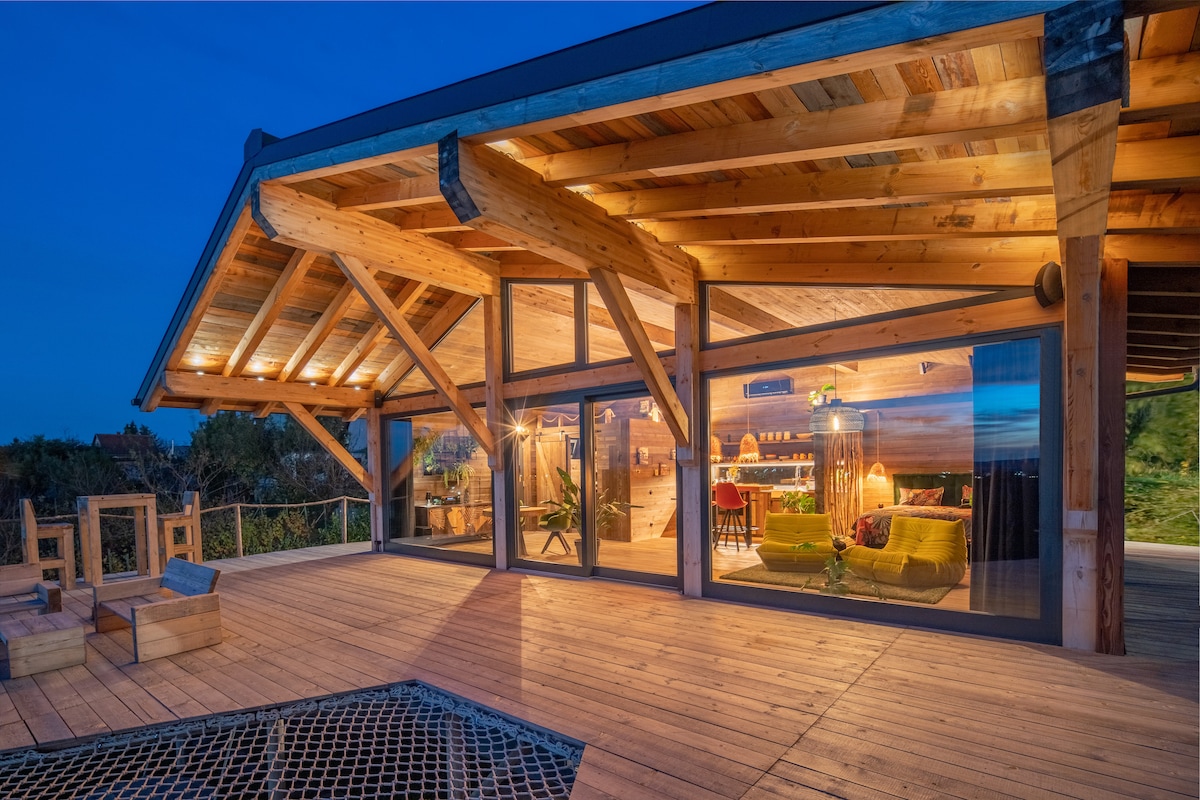 An outdoor wooden deck with a spacious overhang provides shelter. Large glass doors showcase a cozy interior. Soft lighting enhances the warm wood tones, while a glimpse of furnishings and decor can be seen inside. The inviting space overlooks a scenic landscape.