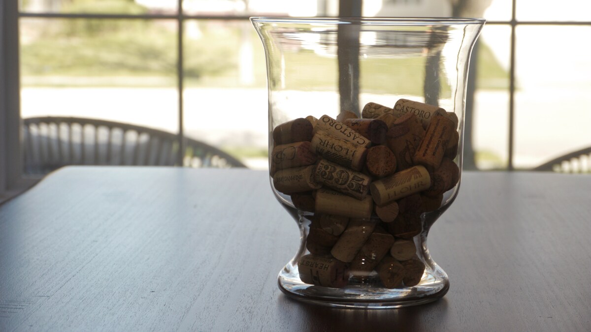 A glass vase filled with assorted wine corks is displayed on a wooden table. Light from a nearby window casts a soft glow, highlighting the textures of the corks. In the background, a view of green grass is visible through the glass panes.