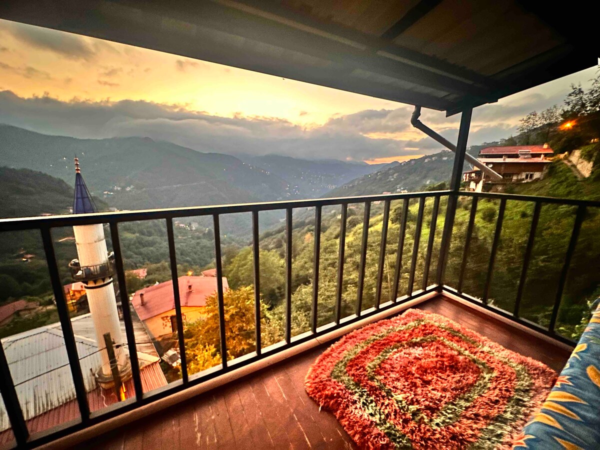 A balcony is presented, featuring a colorful area rug on a wooden floor. Lush green hills are visible in the distance, with a sunset casting a warm glow over the mountains. A peaceful view enhances the outdoor seating experience.