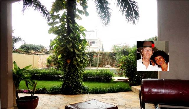 The image showcases a lush garden, framed by tall palm trees and vibrant greenery. A brown couch and a table are visible in the foreground, while a clear pathway leads into the garden, creating a tranquil outdoor space.