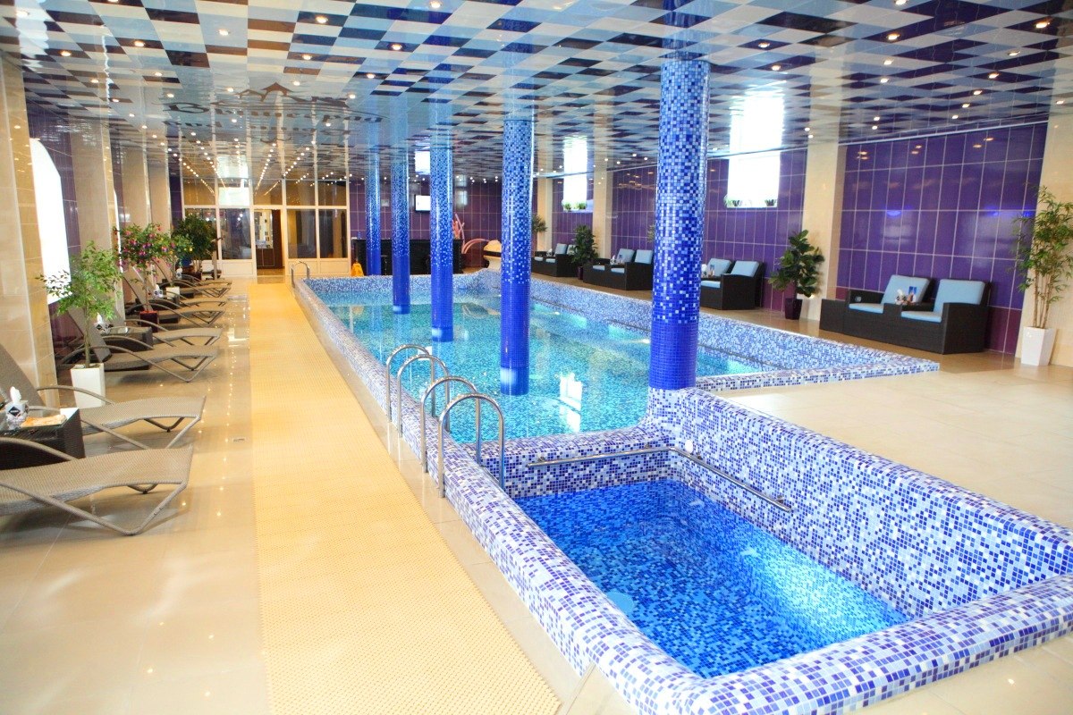 An indoor spa area is depicted, featuring a long swimming pool with mosaic tiles in shades of blue. Several spa lounges line the sides, and tall columns are incorporated into the design. Natural light fills the space, highlighted by decorative plants throughout.