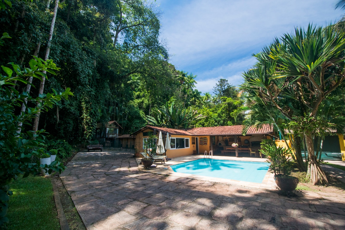 A serene outdoor area features a refreshing pool surrounded by lush greenery. Natural stone tiles lead to the water's edge, while tropical plants frame the space. A shaded seating area is visible nearby, complementing the rustic charm of the yellow building in the background.