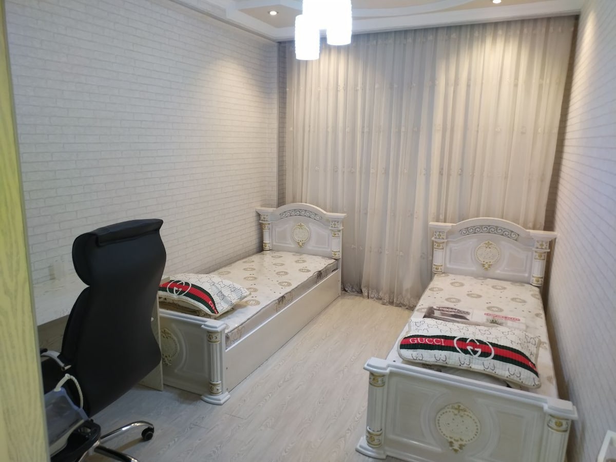 The room features two elegantly designed twin beds with decorative headboards. A sleek office chair is positioned nearby, and sheer curtains allow soft light to filter in. The light-colored walls and flooring create an airy ambiance.