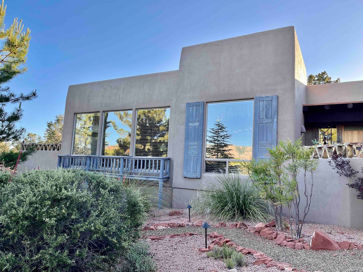 A Southwestern style home features large windows with blue shutters, allowing abundant natural light. Surrounding greenery includes native shrubs and plants, complementing the earthy exterior. The decorative landscape includes gravel pathways, enhancing the tranquil atmosphere of the property.