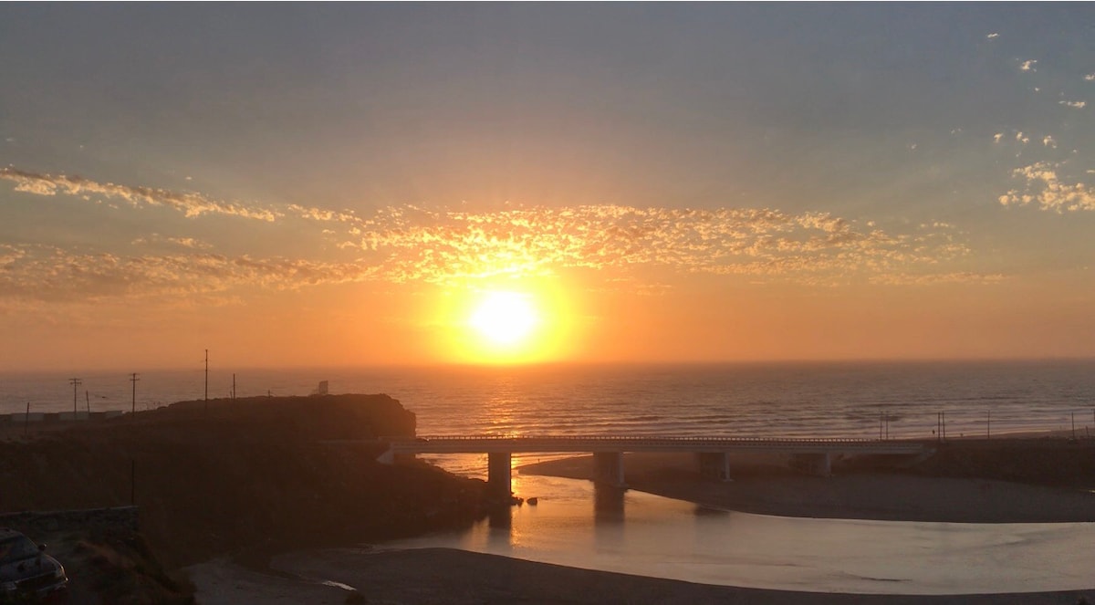 A serene sunset is depicted over the ocean, casting a warm glow across the water. A bridge stretches across the foreground, leading towards a gently illuminated horizon. Soft clouds scatter in the sky, enhancing the tranquil atmosphere.
