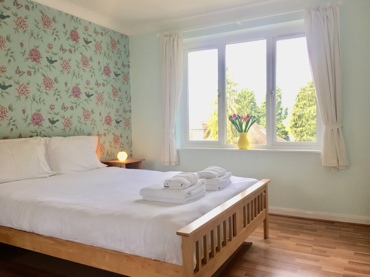 A bright bedroom features a spacious bed with white linens, flanked by two white towels. Floral wallpaper adds a touch of decor, and large windows bring in natural light, offering views of greenery outside. A nightstand with a soft lamp provides additional illumination.